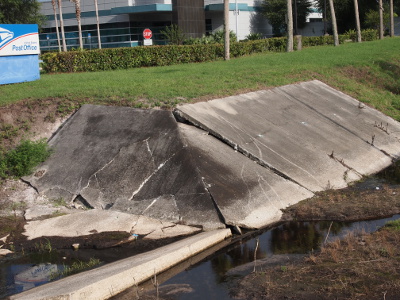 [Several concrete sections cover a corner hillside above the stormwater retention canals. The two sides have rectangular sections while the corner is a triangular section. At the top of the triangle the concrete is lower than the adjoining sides creating an opening which leads under the concrete. There is little water in the canal in this image due to the very dry summer.]